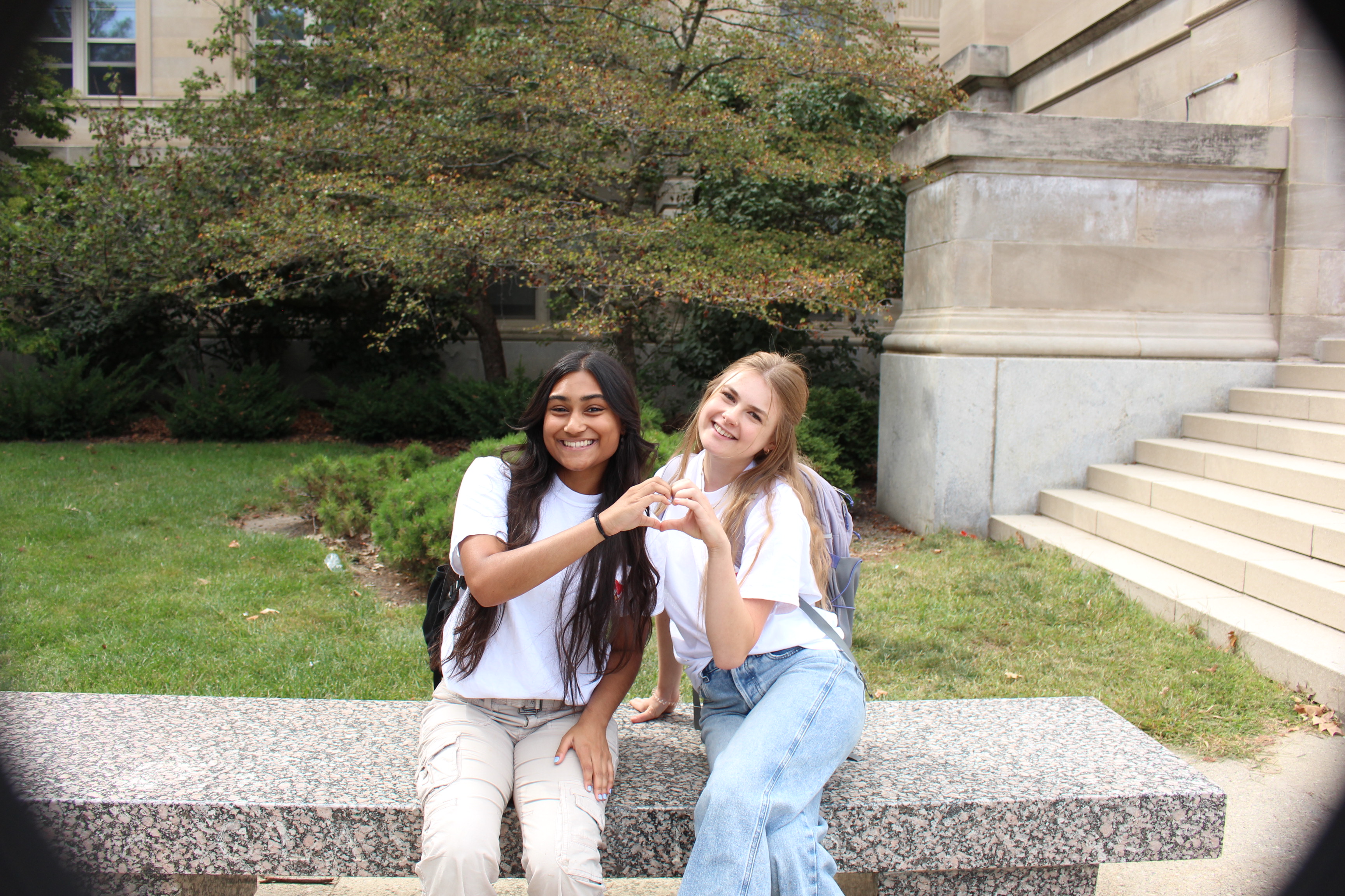 Two students sitting on a bench, smiling and making a heart with their hands.